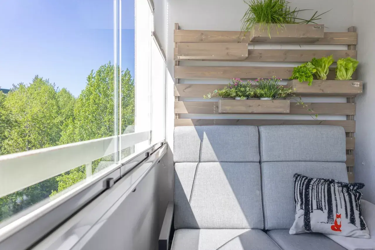 Przytulny mały balkon w bloku z szarą sofą, poduszką z liskiem i zielenią na drewnianej ściance.