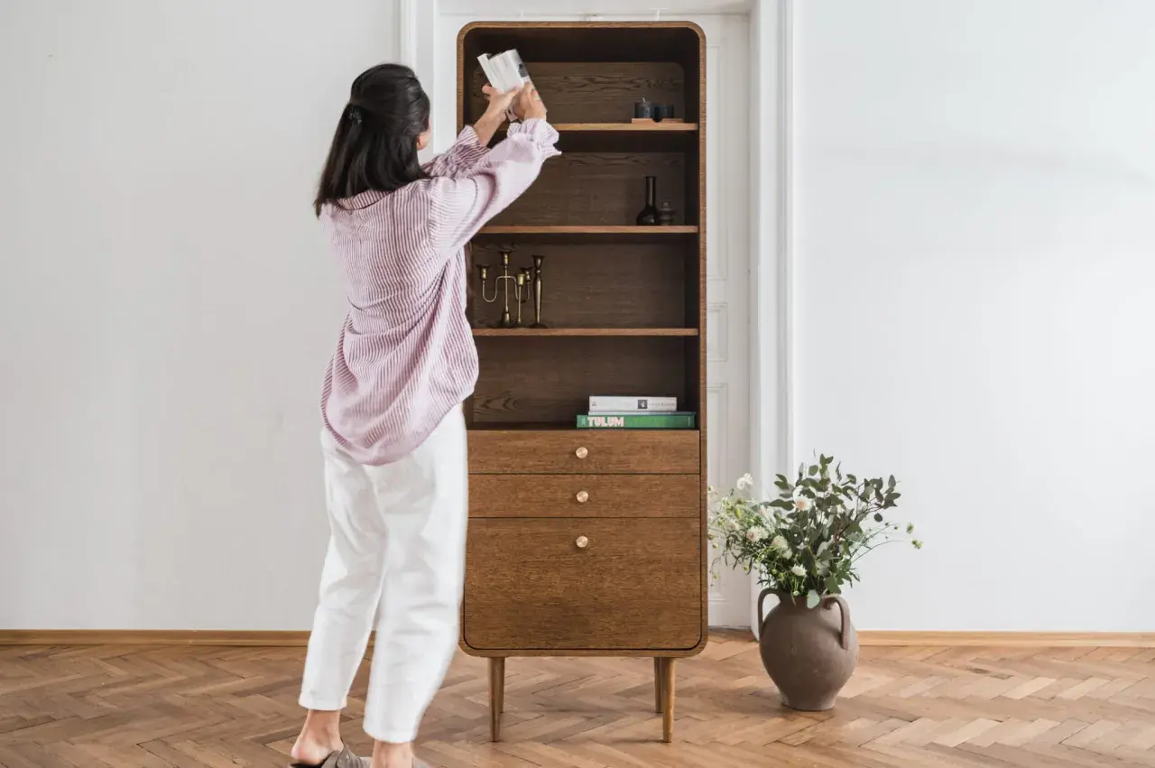 Kobieta układa książki na półce w stylu mid century modern. Drewniany mebel z zaokrąglonymi krawędziami i mosiężnymi uchwytami.