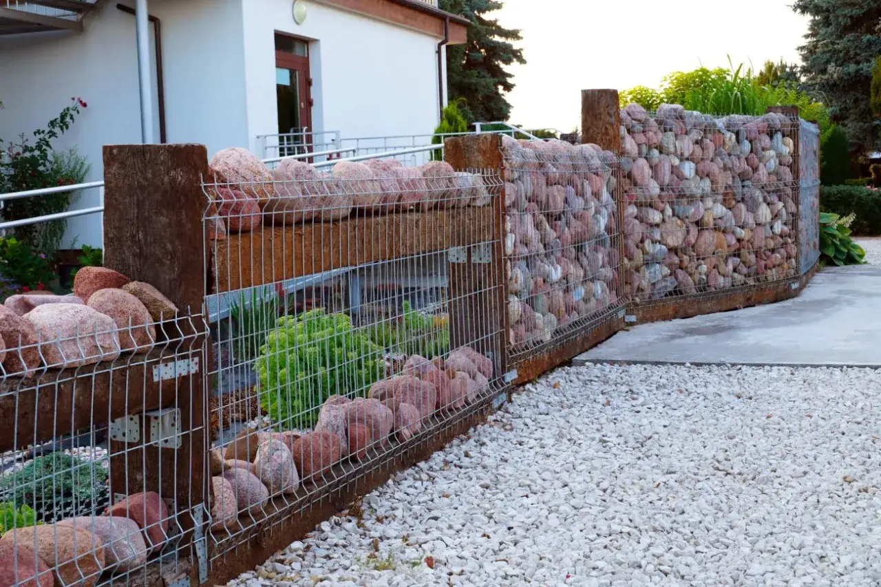 Nowoczesne gabiony w ogrodzie aranżacje: kamienne mury z drewnianymi słupami, otoczone żwirem i zielenią.