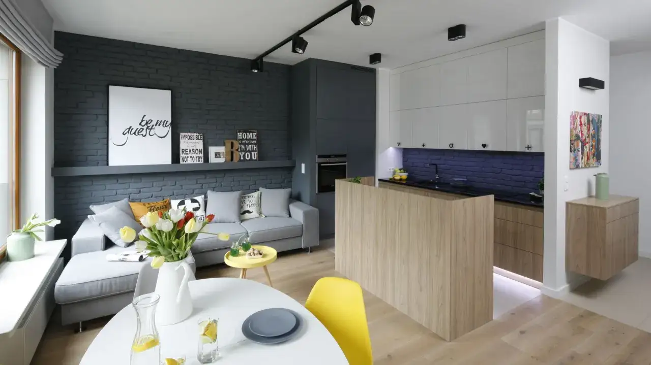 Nowoczesny salon z aneksem kuchennym w bloku. Ceglana ściana, szara sofa i drewniana wyspa kuchenna tworzą przytulny styl loftowy.