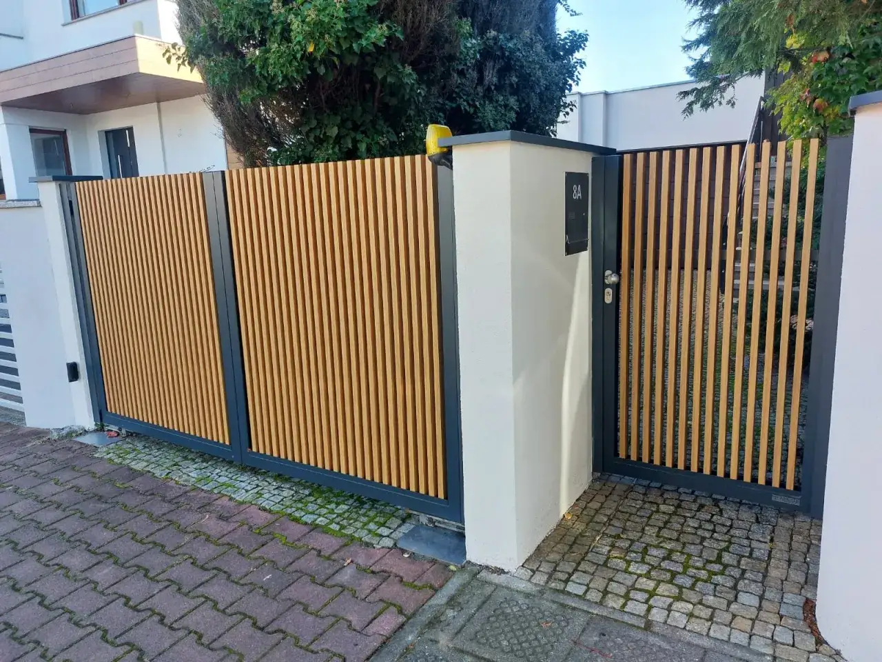 Nowoczesne ogrodzenie przed domem z drewnianymi lamelami i czarną ramą. Brama wjazdowa i furtka tworzą spójną całość.