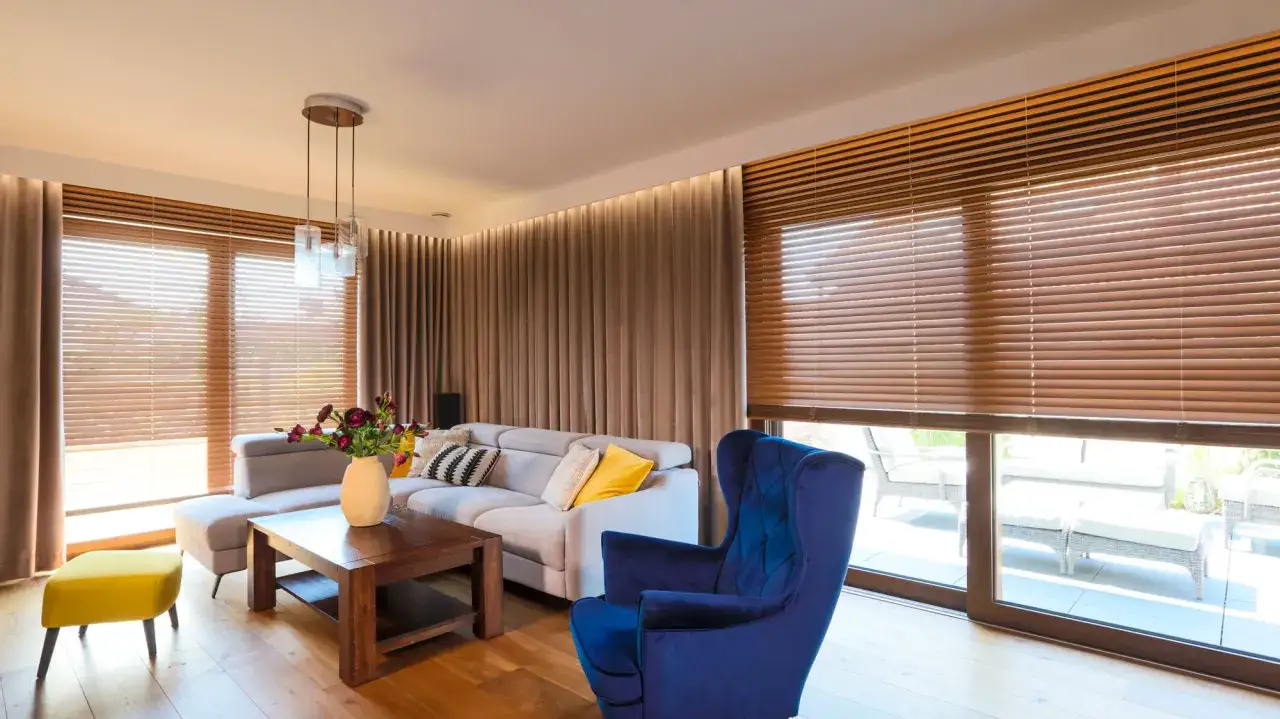 Elegancki salon z szarym narożnikiem, granatowym fotelem i żółtym pufem. Okna zdobią żaluzje drewniane i zasłony, tworząc przytulną atmosferę.