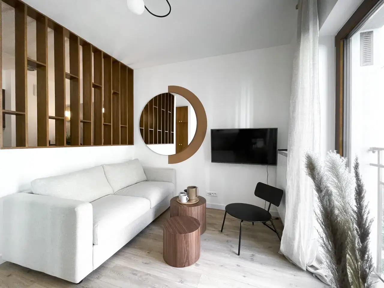 Jasny salon w bloku z sofą, stolikami i telewizorem. Drewniana ażurowa ścianka i okrągłe lustro tworzą harmonię, wprowadzając zasady feng shui w mieszkaniu.