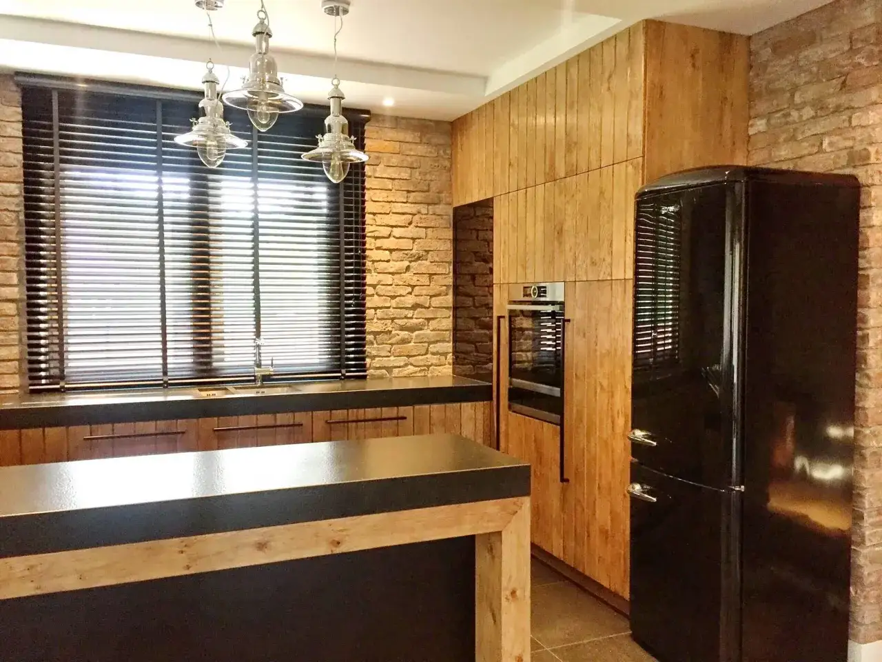 Nowoczesna kuchnia z ceglaną ścianą, drewnianymi frontami, czarną lodówką i wyspą. Okno zasłaniają żaluzje drewniane i zasłony.