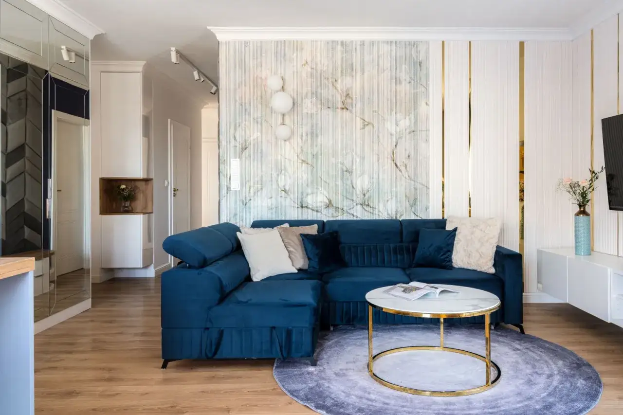 Granatowa sofa i marmurowy stolik tworzą elegancki kącik w salonie. W tle subtelna tapeta i złote detale podkreślają styl glamour.