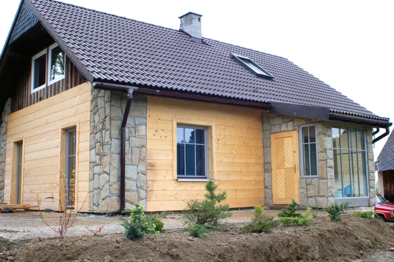 Nowy dom z piaskowca i drewna, z brązowym dachem, oknami i drzwiami. Wokół młode drzewka i ziemia.