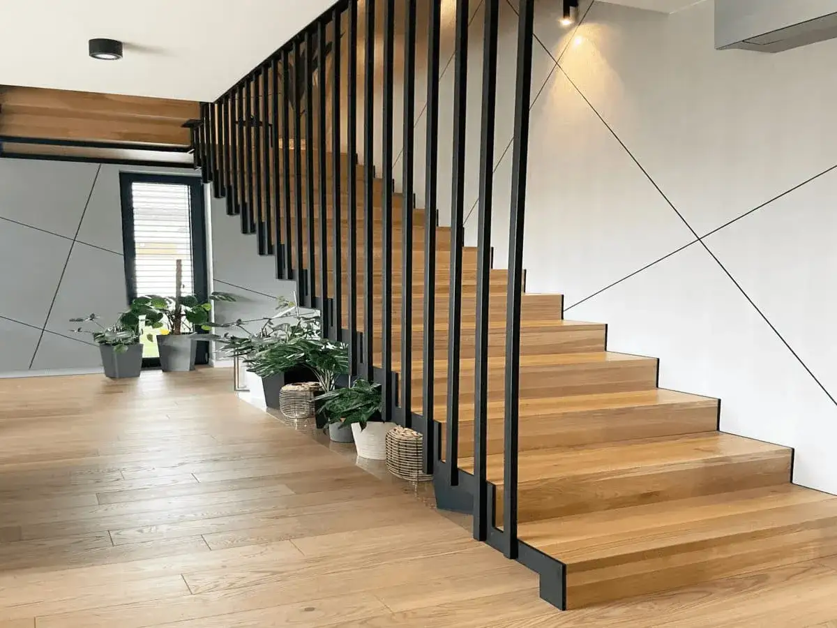 Nowoczesna klatka schodowa w domu z drewnianymi stopniami i czarną, metalową balustradą. Wzdłuż schodów stoją donice z zielonymi roślinami.