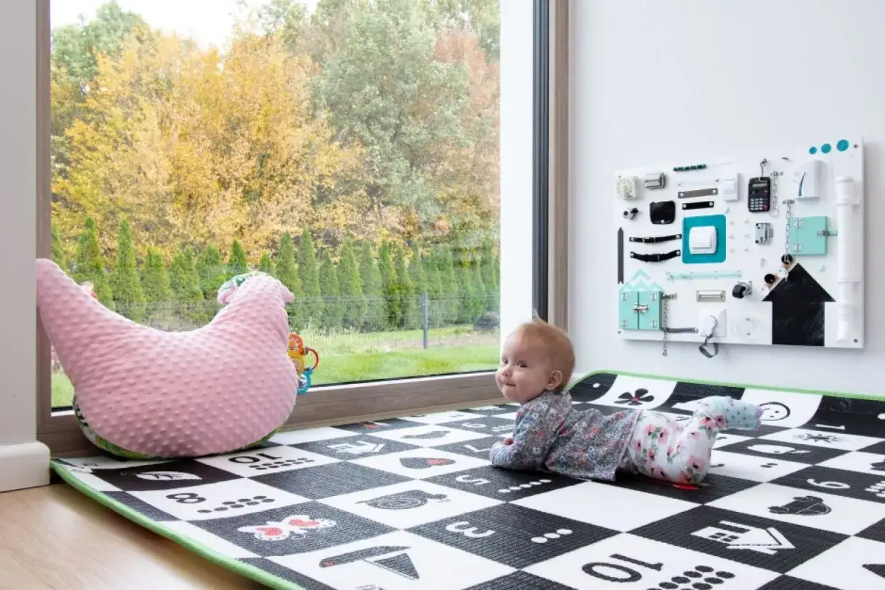 Urocze okno w pokoju dziecka aranżacje: maluch na macie obok różowej poduszki w kształcie kurczaka, za oknem jesienny ogród.