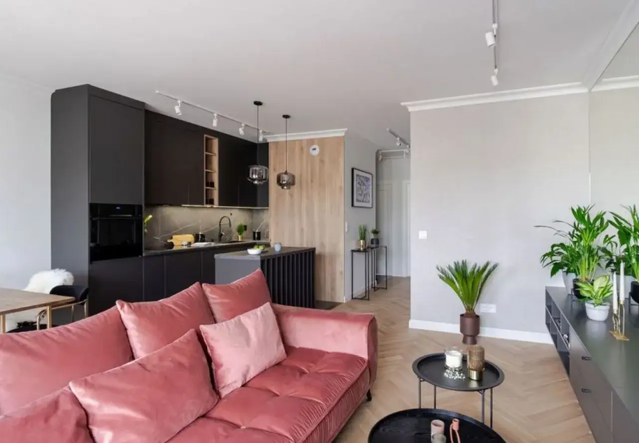 Nowoczesny salon z aneksem kuchennym w projekcie mieszkania 70m2. Różowa kanapa, czarne meble kuchenne i drewniane akcenty tworzą przytulną przestrzeń.