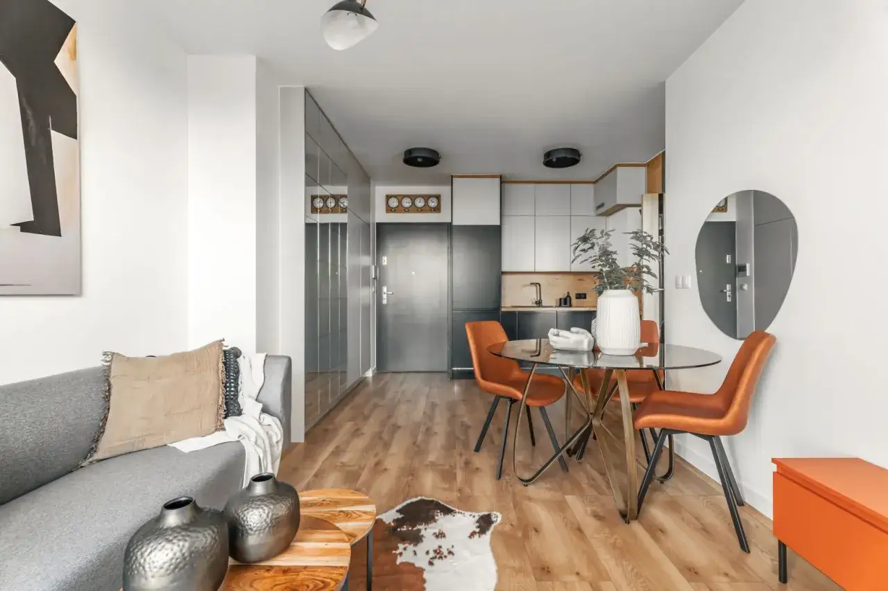 Nowoczesna aranżacja mieszkania 34m2: szara sofa, okrągły stół z pomarańczowymi krzesłami, kuchnia i lustrzana szafa.