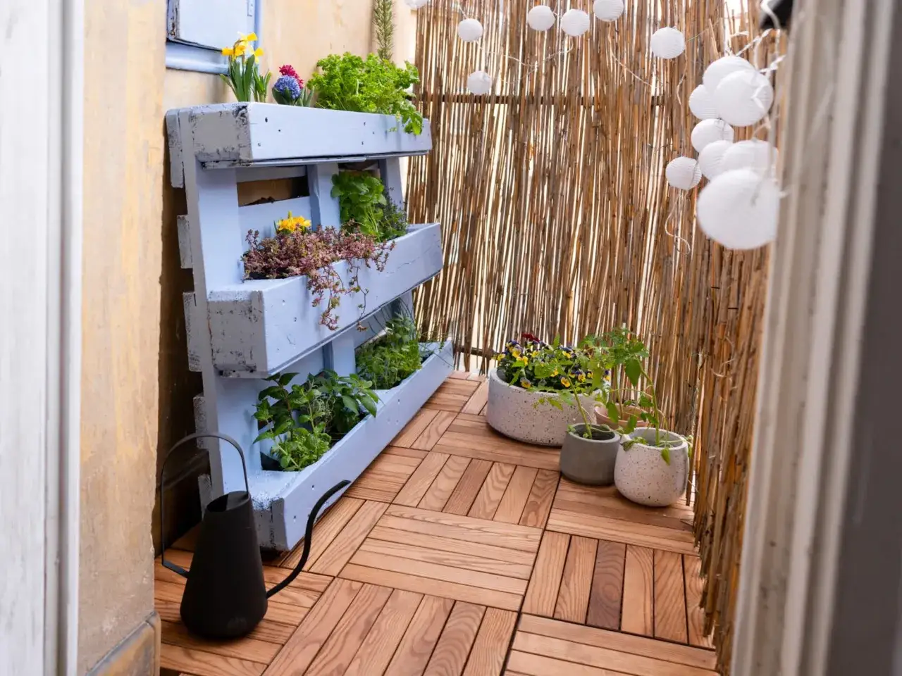Balkon inspiracje: drewniana podłoga, paleta z ziołami i kwiatami, bambusowa osłona i lampiony tworzą przytulną przestrzeń.