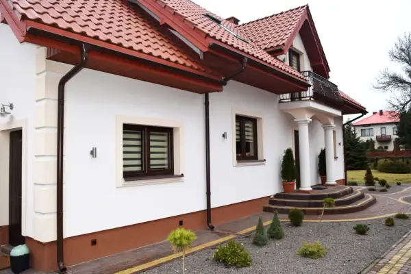 Nowy dom z białymi ścianami i brązowym dachem. Wokół domu widać zadbany ogród z kamiennym podjazdem i ozdobnymi krzewami. Cokół wokół domu jest w kolorze ceglastym.