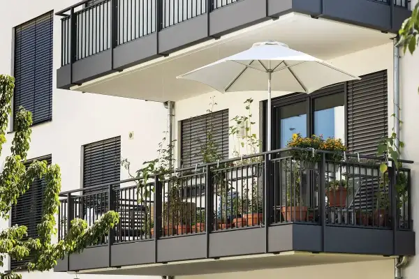 Nowoczesny balkon z białym parasolem i licznymi roślinami w doniczkach. Ciemna balustrada i okna z żaluzjami tworzą elegancką całość.