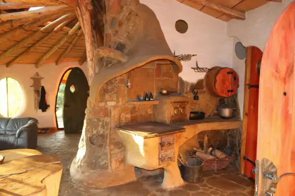 Wnętrze przytulnego domu hobbita z piecem kaflowym, drewnianym sufitem i okrągłymi drzwiami.