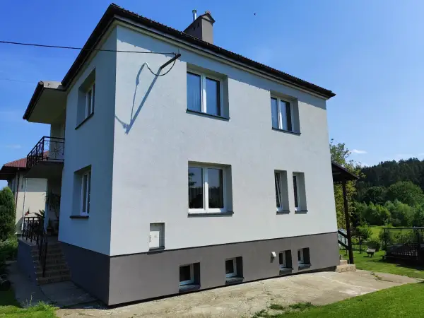 Jasnoszary dom z lat 80. z balkonem i ciemnoszarą podstawą, otoczony zielenią.