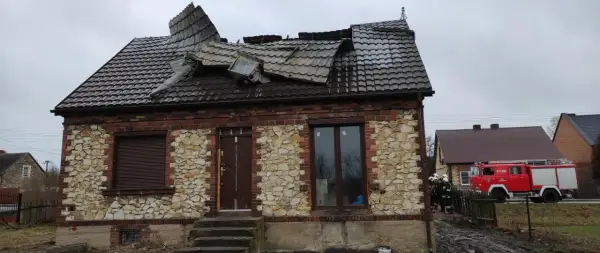 Zniszczony dach domu z kamienia wapiennego. Obok stoi wóz strażacki, a przy nim strażacy.