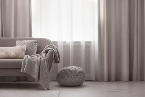 Miękka sofa z poduszkami i pledem, obok okrągła pufa. Elegancka dekoracja okien z zasłonami tworzy przytulną atmosferę.