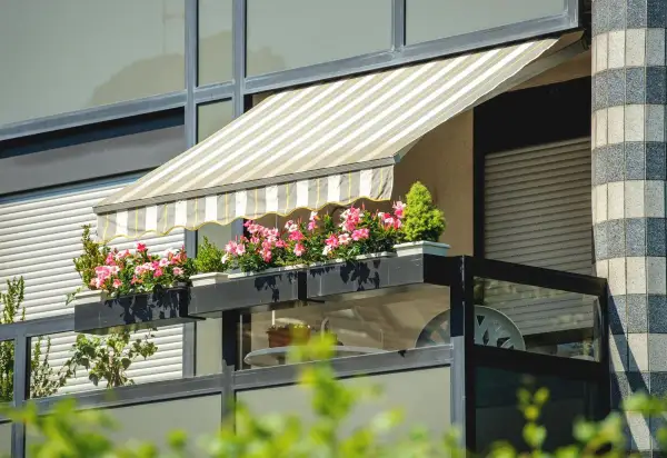 Pasiasty markiza nad balkonem w bloku, z kwitnącymi różowymi kwiatami w skrzynkach, to świetny sposób, jak osłonić balkon.